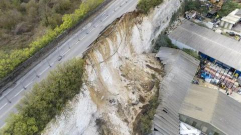 An aerial photo of a collapsed white chalk cliff. Part of the road running along the cliff has collapsed. Below on the right are a number of commercial buildings