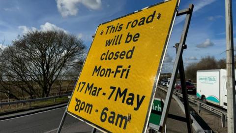 A yellow road sign, stating: This road will be closed Monday to Friday 17 March to 7 May, 8pm to 6am. In the backdrop are trees, a white clouded blue sky, and a queue of traffic including a van bearing the name "Lowestoft Fish Co."