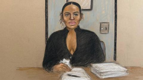 Court sketch of Gabriella Carrington who has pen on a piece of paper next to s stack of papers. She is in front of a blue prison door and is in black
