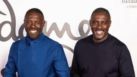 Idris Elba in a black sweater stands next to his lifelike waxfigure in a blue suit.