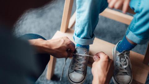 A shadowy figure possibly a woman in black is bending over and tying the shoelaces of a child sitting on steps wearing blue and white pumps. The child is wearing blue socks and jeans and you can only see their legs.