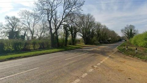 A Google Street View looking at Newington Road, an A-Road, from the Bloxham Road. Tall trees run along one side of the road, a field is beyond.