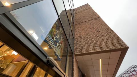 A picture of a modern building with a glass front and a coat of amrs on it.