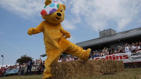 Pudsey bear, a large yellow bear with a polka dot eye patch, jumped over a straw bale with crowds cheering in the background