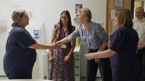 Several hospital staff members gathered in a ward or clinical area. One staff member is shaking hands with Yvette Cooper while colleagues stand nearby. Medical storage units, a doorway and clinical equipment are visible in the background.