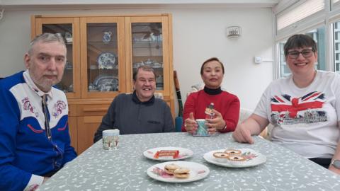 Four people sit around a dining table laden with biscuits and cups of tea.