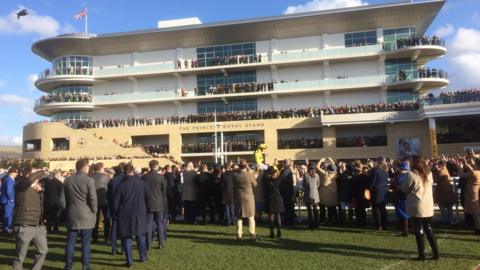 A crowd of men and women standing in front of the Princess Royal stand, looking up at people standing on it. The sky is blue behind the stand and people are dressed smartly, mostly in dark colours, with suit jackets and more formal coats, trousers and shoes. 