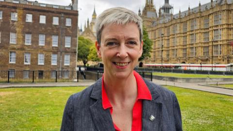 Polly Billington MP is wearing a red shirt and grey jacket. She is standing on Westminster Green with the Houses of Parliament in the background