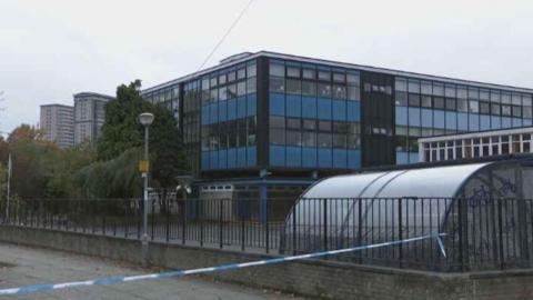 Exterior of a primary school - around three stories high - with a bike shed in the playground and fence around the property, with blue and white police tape sealing off the street in front