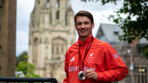 Dan Bethell in Team GB red tracksuit, holding the silver medal he is wearing round his neck, smiling to camera with the Wills Memorial Building out of focus in the background