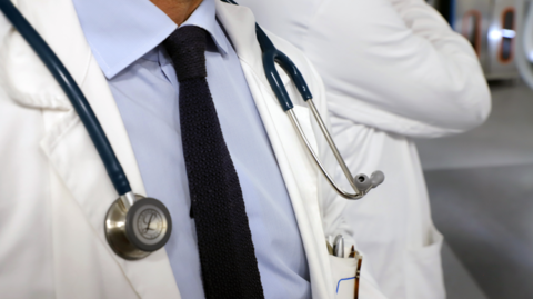 A general view of a doctor in white surgical coat and shirt, with a stethoscope around his neck. The image focuses below chin level