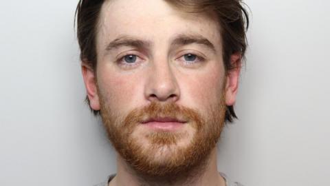 A man with brown/auburn hair and a short ginger beard stands against a grey background