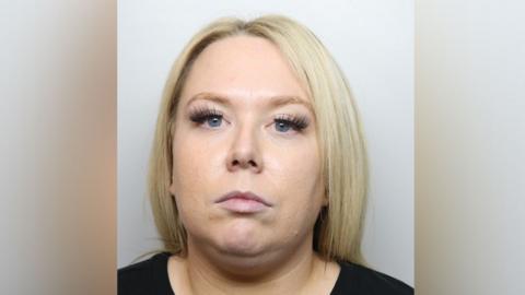 A mugshot of Chelsea Kemp. She has blonde shoulder length straight hair and blue eyes, with false eyelashes. She wears a black top and is staring at the camera. 