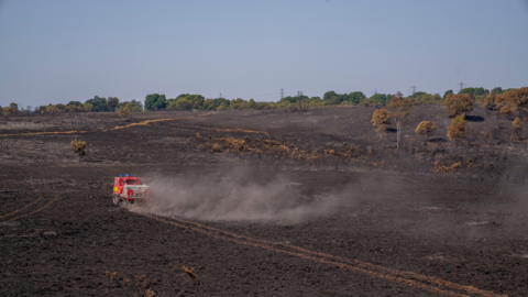 A picture that shows large areas of damage after the fire and a fire service vehicle driving across the scorched land.