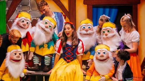 A female performer on stage dressed in bright yellow skirt, a blue bodice with red detail and puffed white sleeves. She is surrounded by seven other performers holding puppets with white beards and yellow hats. Their costumes include colourful tunics paired with brown shorts and striped socks and dark shoes.