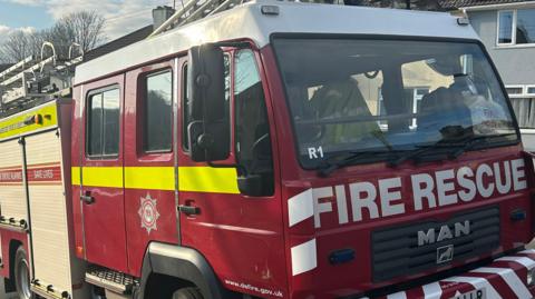 A red Devon and Somerset Fire Service engine at the scene of an incident in a residential area. No-one is sitting inside the vehicle. The engine is made by MAN and has fire rescue written in white block capitals on the front. The fire service's badge is on the side of the vehicle.