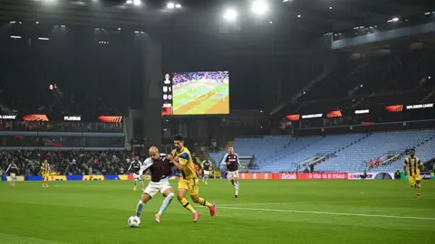 Footballers play on the pitch with empty away stand in the background. An electronic screen shows the score as 1-0