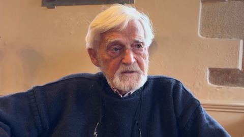 A man sits in front of a white stone wall. He has a white beard and white hair. He is wearing a navy blue jumper with a shirt underneath.