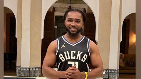 Person in a black Milwaukee Bucks  jersey and shorts standing indoors before ornate tiled arches and a decorative fountain 