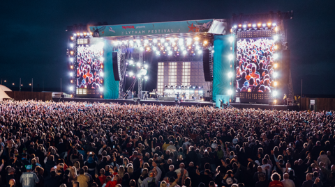 A wide shot of thousands of people watching an act at Lytham Festival in the evening. The stage is lit up with 'Lytham Festival' at the top and two large screens to the side showing the crowd.