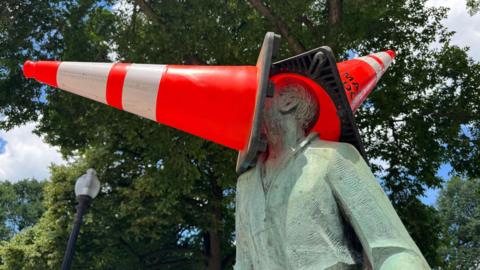 A person in a park wearing a blue hat and a sleeveless sports jersey with the number 23 places a bright orange traffic cone on top of a seated statue’s head, which is already covered by a larger cone. The statue sits on a decorative base surrounded by grass and trees, with city buildings visible in the background on a sunny day.