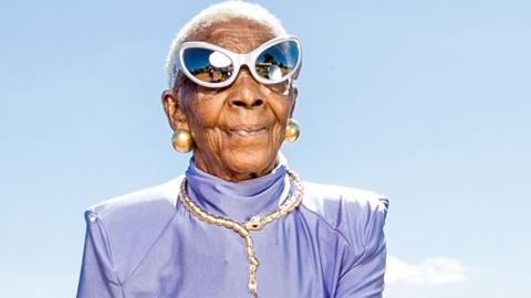 Margret Chola in a shiny lilac top wearing cat-eye mirrored sunglasses, a bejewelled gold necklace  in the shape of a snake and big gold ball earrings