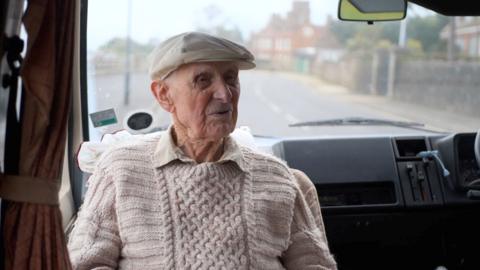 Bill wearing a cream cap and a beige jumper and shirt sat inside a van with the road behind him