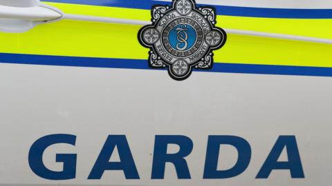 The side of a white and yellow Garda car, the Garda symbol is in shot along with 'Garda' written in blue writing.