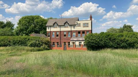 A Victorian three-storey property with a sloping roof stands flanked by large hedgerows in the middle of a grass field.