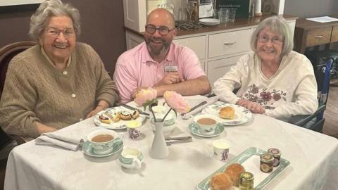 A square table covered in a white table cloth, with a tray of jam, cream and scones, tea cups containing tea and plates with scones on. Betty is sitting to the left with medium-length grey hair and a brown cardigan, Martin has very short hair and a brown beard and glasses and is wearing a pink shirt, and Jean has medium-length white hair and is wearing a white cardigan with pink flowers.