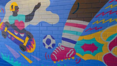 A mural representation of a skateboard being ridded by a child like figure, and a leg with a football boot and a football being kicked. 