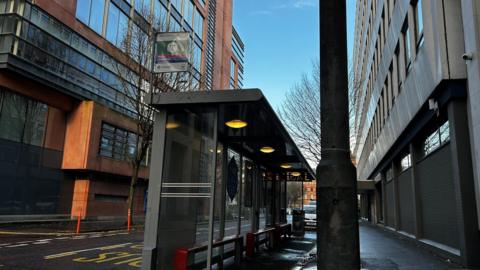 A day time picture showing a bus stop and shelter. The Translink sign attached to the shelter says Adelaide Street. Two large buildings with lots of windows can be seen either side of the street. A bare tree can be seen behind the shelter. A lamp post is to the right of the picture in the foreground.