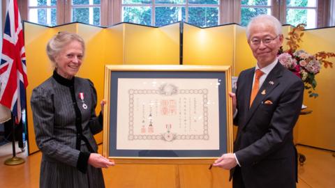 Jane Hasell-McCosh on one side, with a medal on her jacket, holding a framed certificate in Japanese, with Hajime Hayashi on the other side wearing a suit and orange tie, also holding the frame.