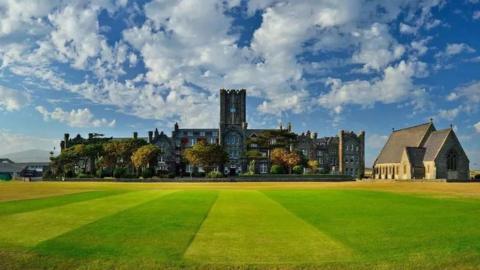 A neatly mown green lawn in front of a large grey building with a tall clock tower in the middle. A chapel building sits to the right.