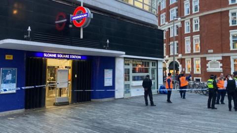 Sloane Square Tube station with blue police tape blocking the entrance as staff and police officers stand to the right, some wearing fluorescent tabards