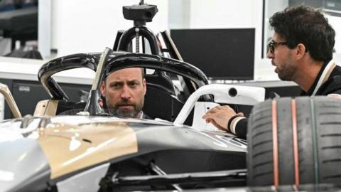 The Prince of Wales sits inside a car during a visit to Formula E Team Jaguar TCS Racing in Kidlington. 2020 world champion Antonio Felix da Costa is besides the car, speaking to him. Two more men are behind them, smiling.