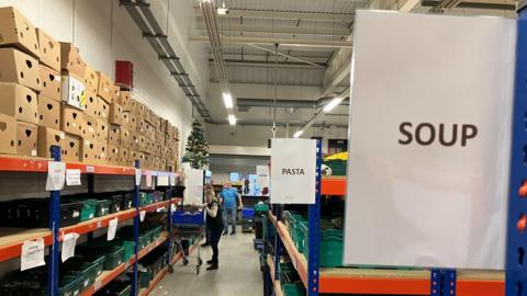 A long aisle in a warehouse stacked with boxes and cans of food. There are signs saying "Pasta" and "Soup". A woman with a trolley is in the distance picking up cans.
