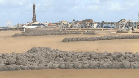 Visualisation of rock headlands along the beach. They are long stretches of grey rocks edging from the steps to the beach towards the sea. Blackpool's coastline can be seen in the distance in the background with the tower to the left