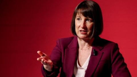 Rachel Reeves, a woman with a dark brown bob, wearing a burgundy jacket and white blouse, gestures with her right hand as she stands against a red background