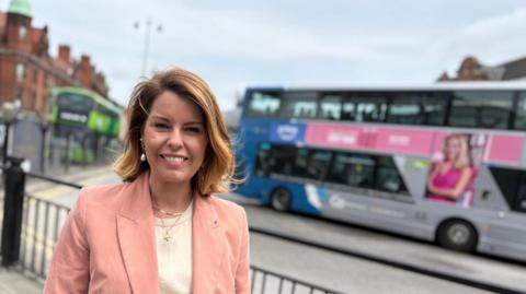 North East mayor Kim McGuinness at Haymarket bus station in Newcastle city centre. She is wearing a light pink jacket and smiling at the camera. There are two double decker buses behind her, out of focus.