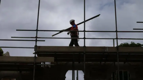 A construction worker in silhouette carries scaffolding poles over his shoulder on a building site