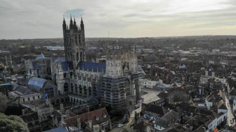 Canterbury Cathedral, which is a large, stone cathedral in gothic style. It is partly covered with scaffolding and towers over the surrounding city of Canterbury.