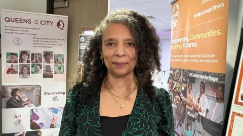 Jennifer Blake, a "Queen" of Walsall, at the Queens of the City event by LGK in the Community. She has long black curly hair past her shoulders and wears a black and green patterend jacket over a black top. Around her neck are two gold chain necklaces.