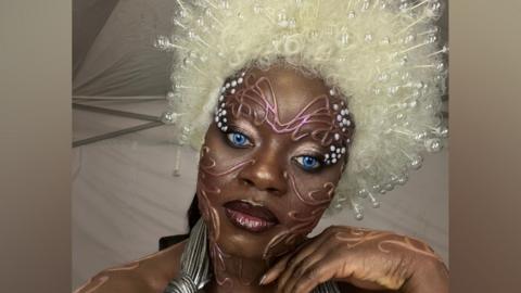 A person in bold avant‑garde makeup with bright blue eyes, a large white beaded wig, and metallic costume accessories, standing in a tent-like backstage setting
