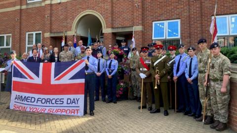 Members of the armed forces gathered at the flag-raising ceremony in Grantham. The personnel are standing behind a union flag with the wording "Armed Forces Day - Show Your Support". They are standing in front of a large red-brick building with arched architectural features and a crest mounted above the entrance.