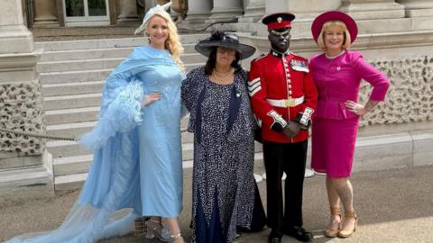 Three women, in formal dresses, standing next to a palace guard wearing a red coat.