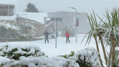 Two people ski along a snow-covered road during heavy snowfall