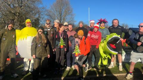 A group of people standing in a line, some wearing fancy dress, smiling, on a sunny day 