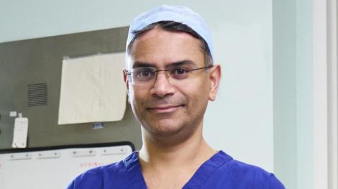 Animesh Patel is wearing a blue medical uniform with a white hat. He has short brown and grey hair and is wearing glasses. Behind him is a wall with a pin board.
