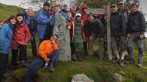 Mark Wood explorer with a large group of teachers and pupils of Sherbourne Fields Special School, they are standing next to a wooden sign that says Snowdown Summit